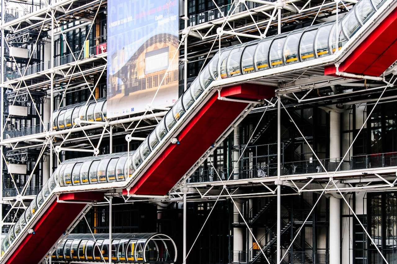 The image shows the exterior of the Centre Pompidou in Paris, characterized by its high-tech architectural style. Prominent features include external escalators encased in transparent tubes and exposed structural elements in a lattice pattern.