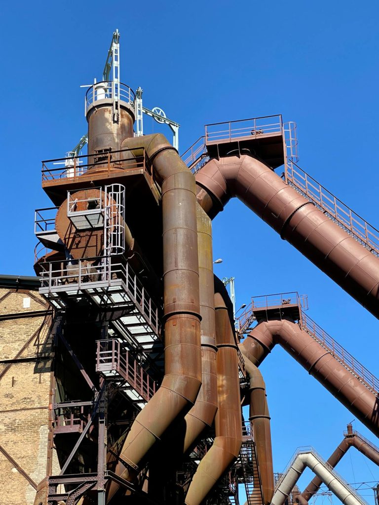 A close-up view of a rusted industrial structure against a clear blue sky. The structure features interconnected pipes, metal platforms, and ladders, giving it a complex and towering appearance.