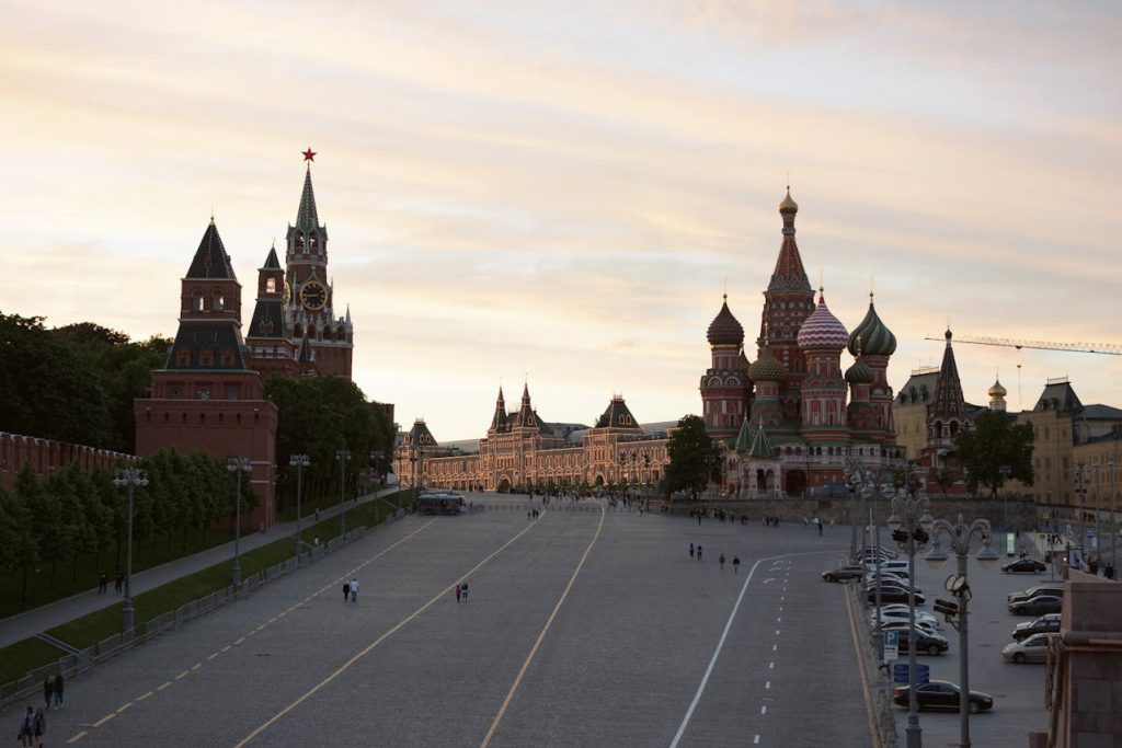 View of Red Square in Moscow at dusk. The image features the Spasskaya Tower on the left with its prominent clock, and the iconic colorful domes of St. Basil's Cathedral on the right. Several people are walking on the square, and cars are parked on the side.