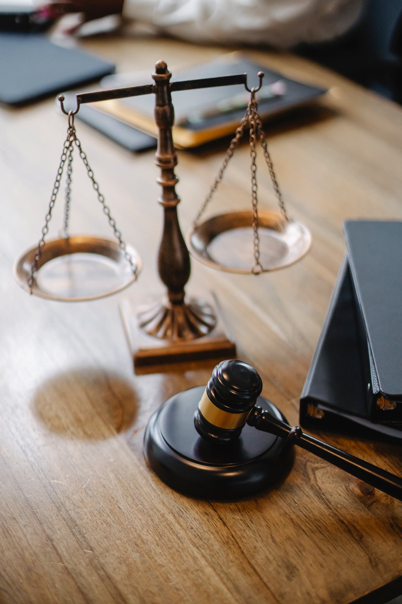 A bronze balance scale and a wooden gavel rest on a wooden table. The background includes some blurred black binders, suggesting a legal or courtroom setting.