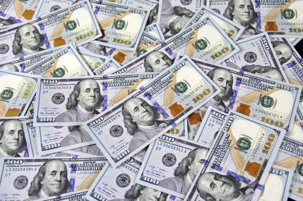 A large pile of overlapping U.S. one-hundred-dollar bills. The notes feature a portrait of Benjamin Franklin and contain blue security threads, green and gold printing, and various intricate patterns denoting their authenticity.