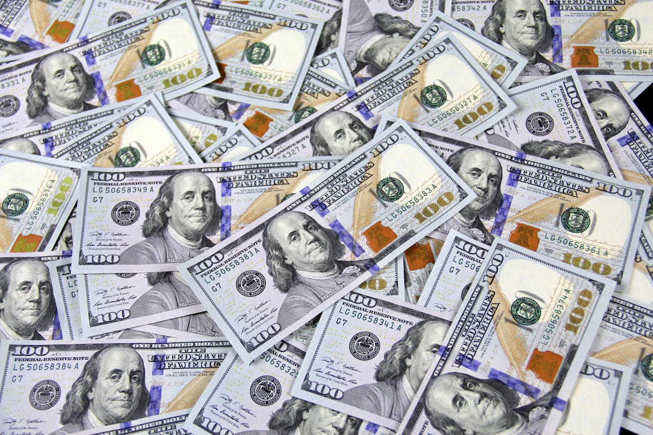 A large pile of overlapping U.S. one-hundred-dollar bills. The notes feature a portrait of Benjamin Franklin and contain blue security threads, green and gold printing, and various intricate patterns denoting their authenticity.