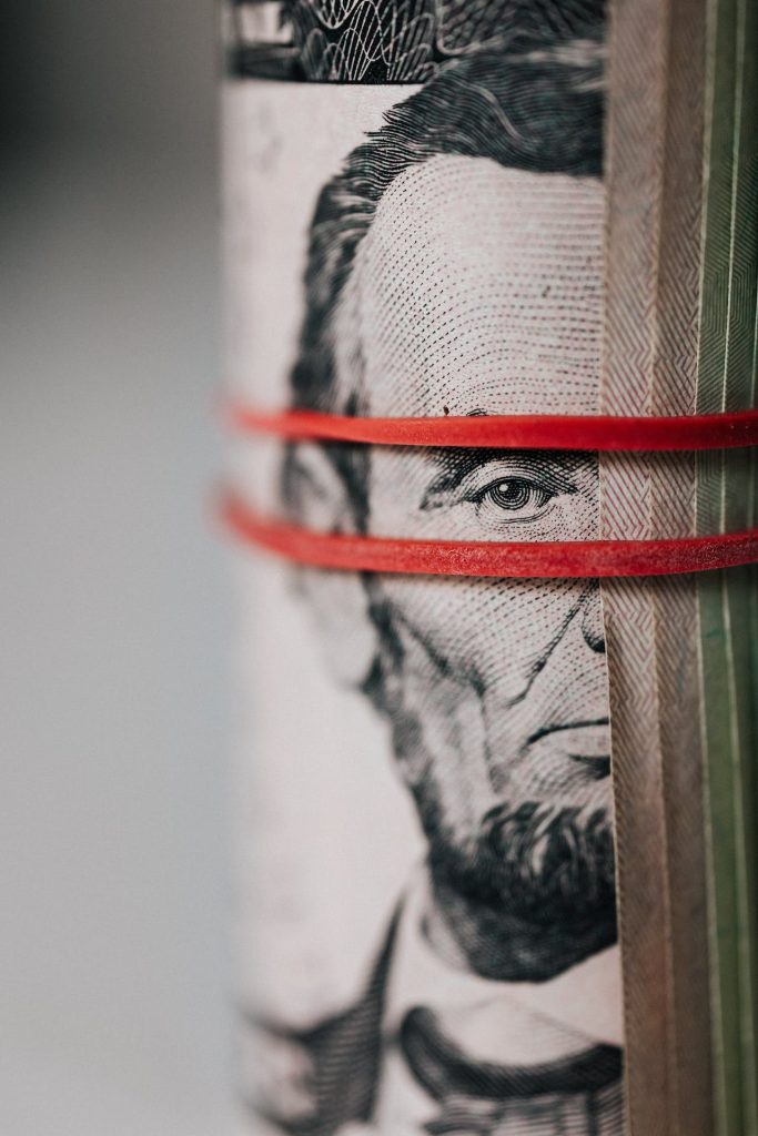 Close-up of a rolled U.S. five-dollar bill with a portrait of Abraham Lincoln visible. A red rubber band is wrapped around the bill, securing it tightly. The background is blurred, drawing focus to the intricate details of Lincoln's face on the bill.