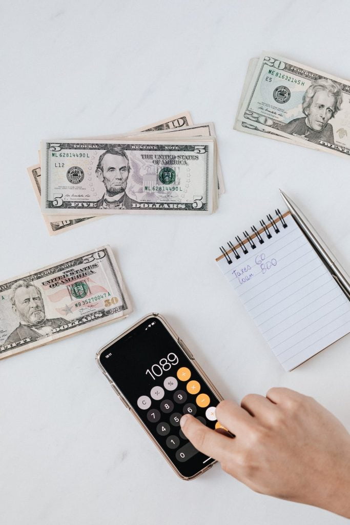 A person is using a calculator app on a smartphone, showing the number 1089. Nearby, there are stacks of U.S. dollar bills and a notepad with a handwritten note that reads "Taxes: 60, Loan: 820." The scene is set on a white surface.