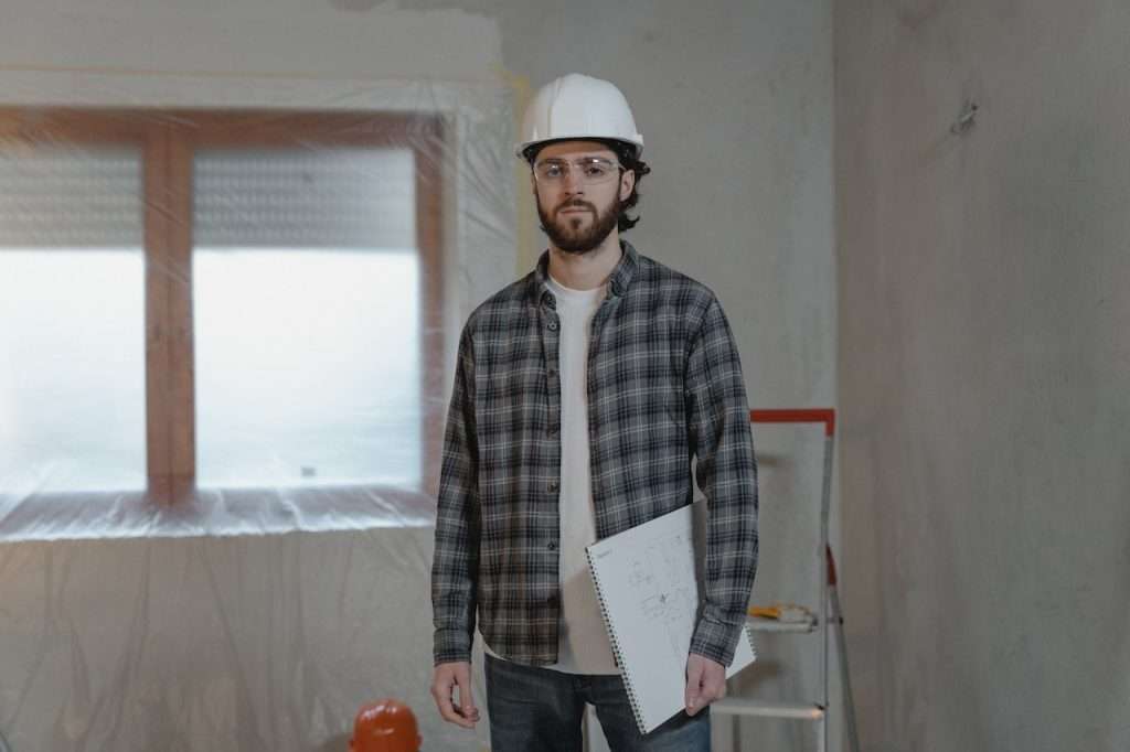 A man wearing a hard hat, safety goggles, and a plaid shirt is holding a notebook with sketches. He stands in a partially renovated room with plastic covering the window. A ladder and various construction tools are visible in the background.