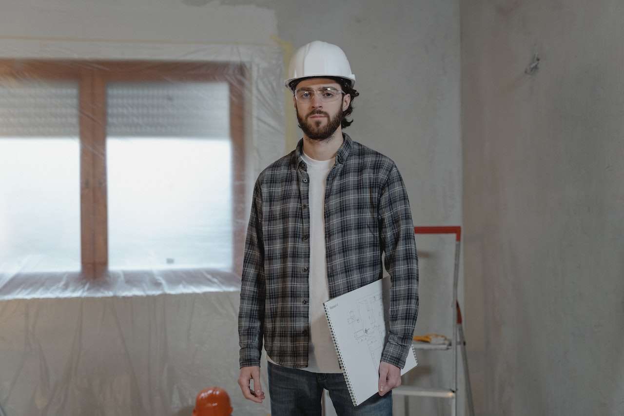 A man wearing a hard hat, safety goggles, and a plaid shirt is holding a notebook with sketches. He stands in a partially renovated room with plastic covering the window. A ladder and various construction tools are visible in the background.