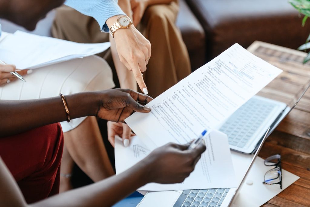Close-up of diverse hands holding and pointing at a document during a meeting. The document appears to be a resume or report, and laptops are visible on the wooden table. The atmosphere suggests a collaborative work environment.