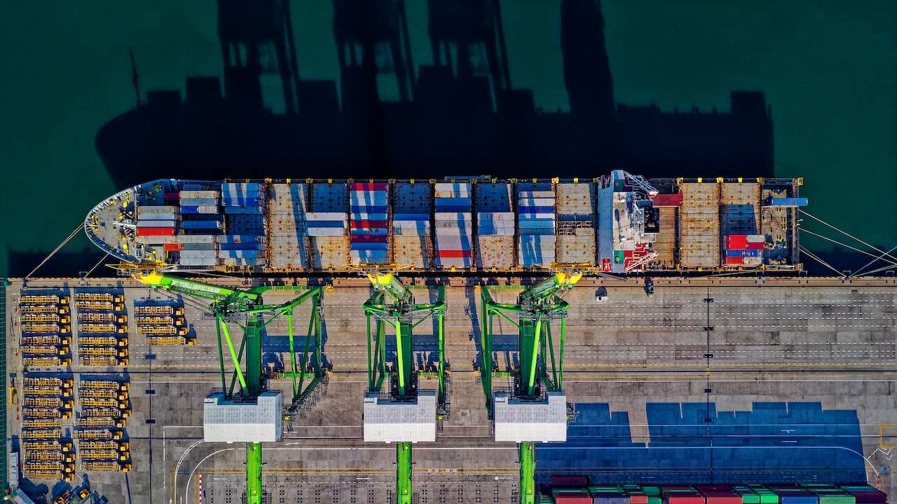 Aerial view of a cargo ship docked at a port. The ship is loaded with multicolored shipping containers. Three large green cranes are positioned alongside the ship, with their shadows cast on the deck and water. The port area has rows of containers.