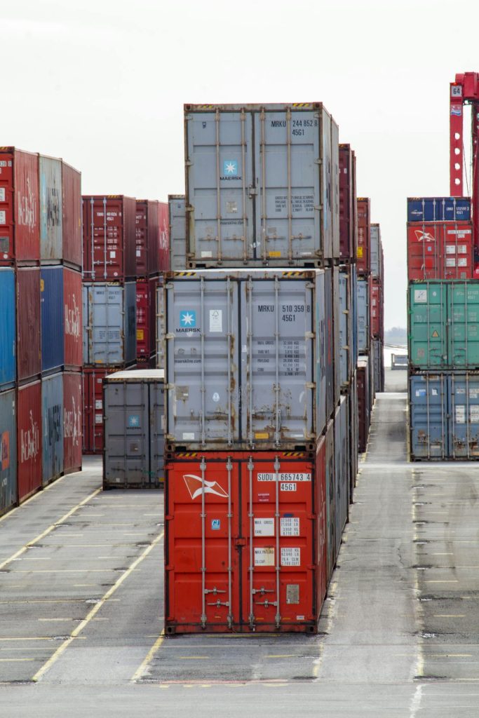 A large stack of colorful shipping containers arranged in rows at a shipping port. The containers are of various colors and marked with different company logos and identification numbers. The concrete ground between the stacks is clear of any vehicles or equipment.