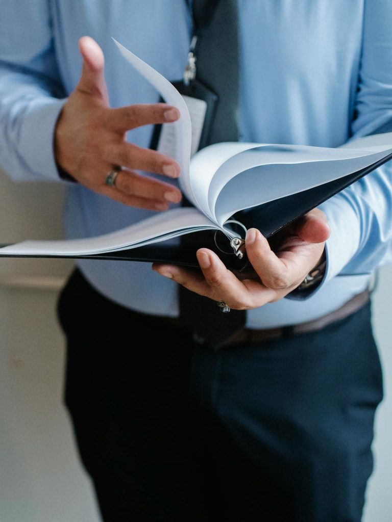 A person wearing a blue shirt and a dark tie flips through a notebook or binder. The individual's hands are in focus, with one hand holding the book open and the other turning a page. The image emphasizes the act of reading or presenting information.