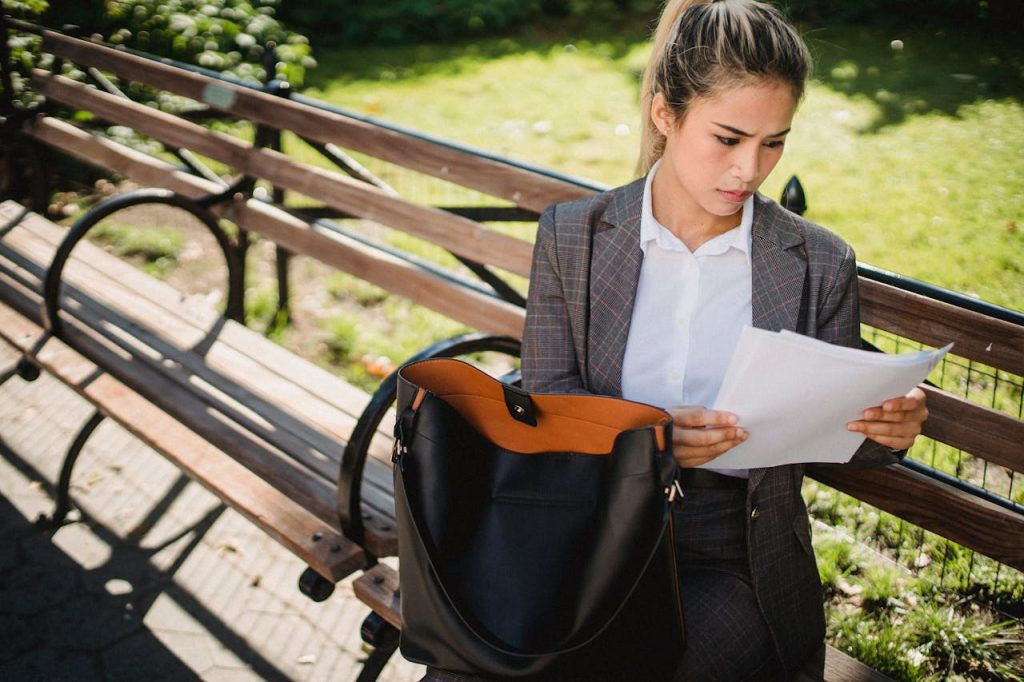 A woman in a business suit is sitting on a park bench, intently reading a document. Her black handbag is open beside her on the bench. The background features greenery and another empty bench.