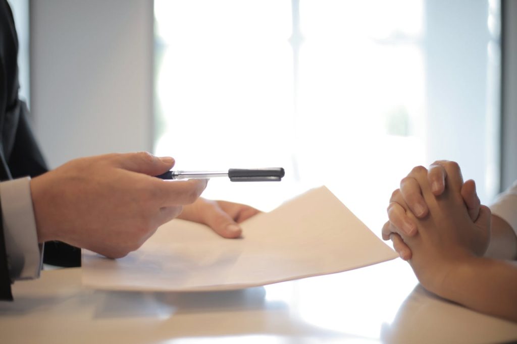 Close-up of two people sitting across from each other at a table. One person is handing a pen and a document to the other, who has clasped hands resting on the table. The background is bright and blurred, suggesting a well-lit room or an office setting.