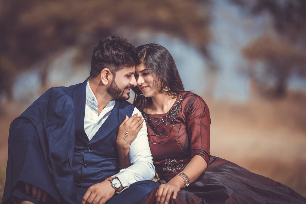 A couple is sitting closely together outdoors, looking at each other with smiles. The man is wearing a blue suit and white shirt, while the woman is in a maroon dress with intricate patterns. The background is blurred, focusing attention on the affectionate pair.