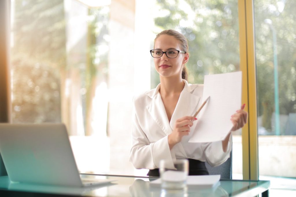 A woman wearing glasses and a white blouse sits at a desk with a laptop and a glass of water. She holds up a piece of paper and points to it with a pen, smiling slightly. The background shows large windows with a blurred view of trees outside.
