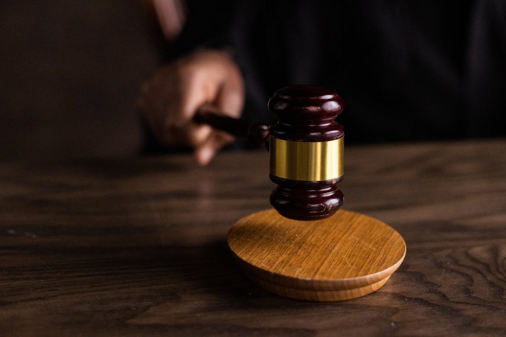 A judge's hand holding a wooden gavel with a brass band over a wooden sound block on a dark wooden table, ready to strike. The image captures the moment before the gavel is brought down.