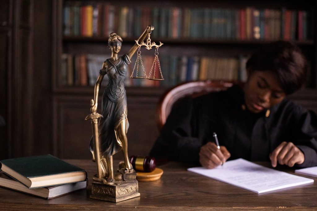 A bronze statue of Lady Justice holding scales and a sword is on a desk with books and a gavel. In the background, a person in judicial robes is writing on a document. Bookshelves filled with books are visible in the background.