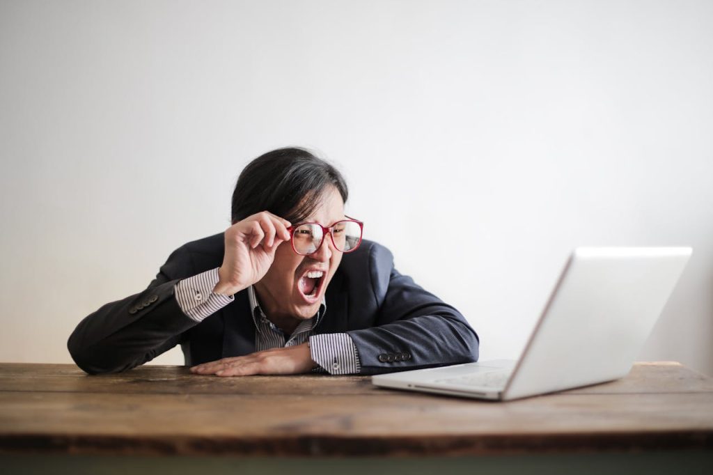 A person wearing a suit and glasses, with long hair, appears surprised or frustrated while looking at a laptop screen on a wooden table. The background is plain and white.