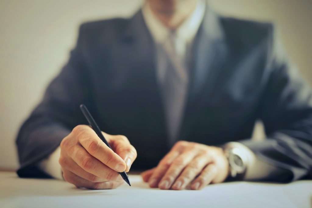 A person in a suit sits at a desk, writing with a pen on a piece of paper. The image focuses on their hands and the pen, with their face and upper body blurred in the background. The setting appears to be a professional or formal environment.