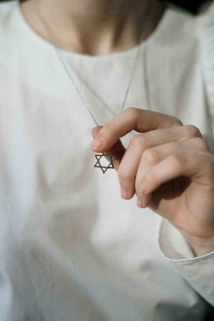 A person wearing a white shirt is holding a necklace with a Star of David pendant. The person's face is not visible in the image.