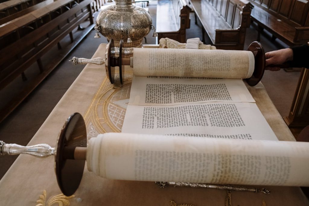 A person is adjusting an open Torah scroll on a table in a synagogue. The Torah scroll is unfurled, displaying Hebrew text. The table is covered with an ornate cloth, and silver ceremonial items are present. Wooden pews are visible in the background.