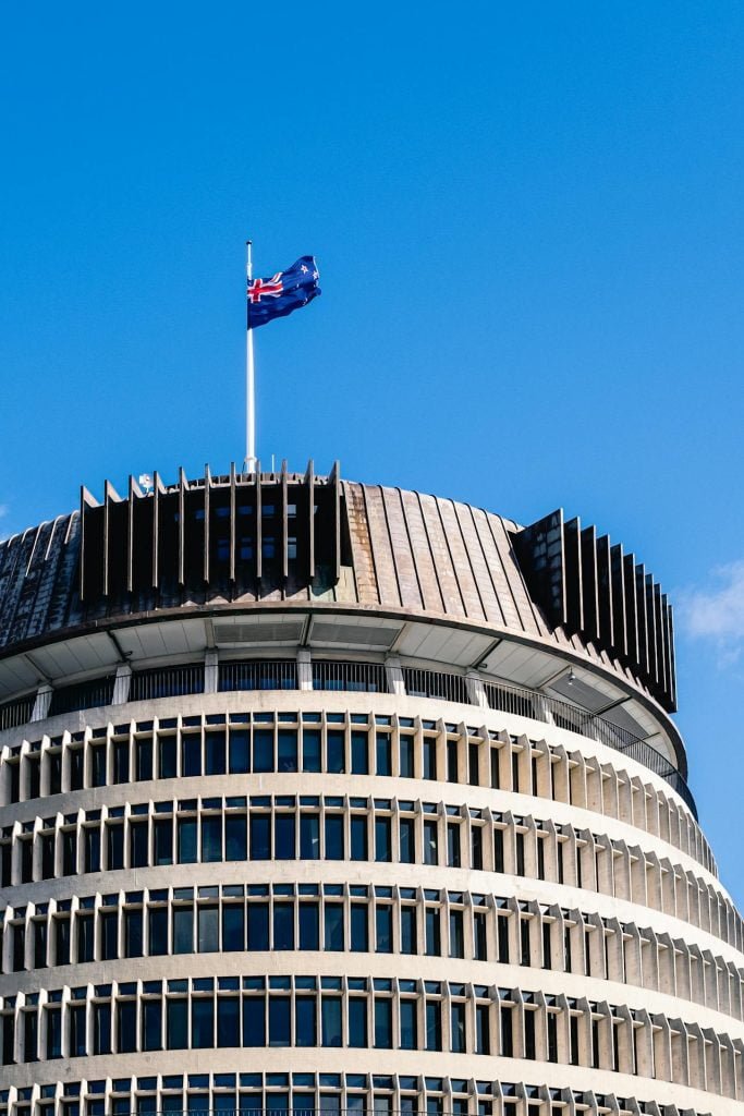 A cylindrical building with a unique, modern architectural design, featuring rows of horizontal windows and a flagpole on top with a blue flag bearing a red and white design, set against a clear blue sky.