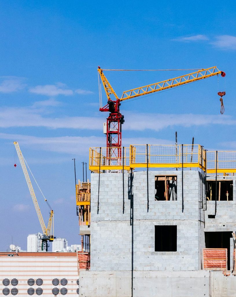 A construction site under a clear blue sky, featuring a partially built multi-story building with scaffolding and cranes. The bright yellow construction crane towers over the structure, while another crane is visible in the background.