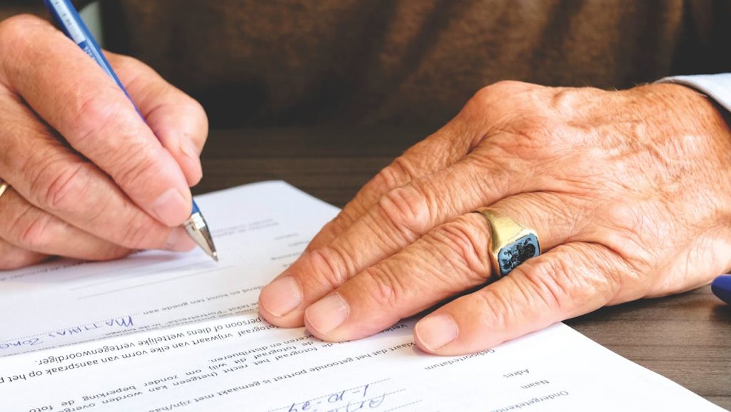 Close-up of an elderly person's hands signing a document. The left hand holds a pen, while the right hand, adorned with a ring, steadies the paper on a wooden table. The document is partially visible with printed text on it.