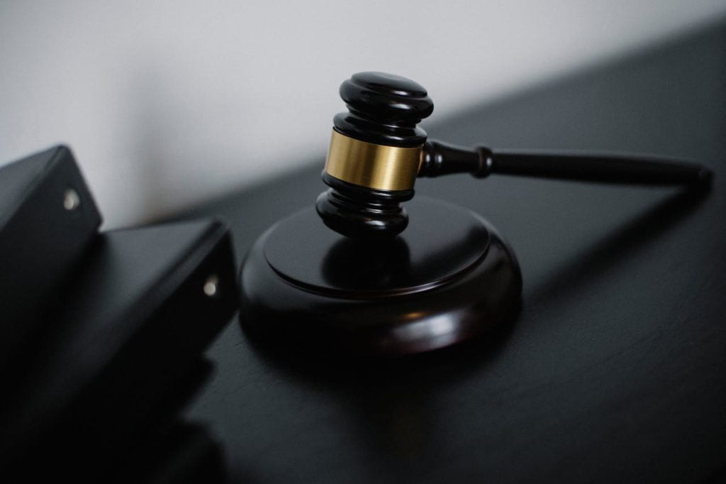 A close-up image of a dark wooden gavel with a brass band, positioned on a matching sound block. Part of the gavel handle is visible. Three black folders or binders are slightly blurred in the background. The setting appears to be a desk or table.