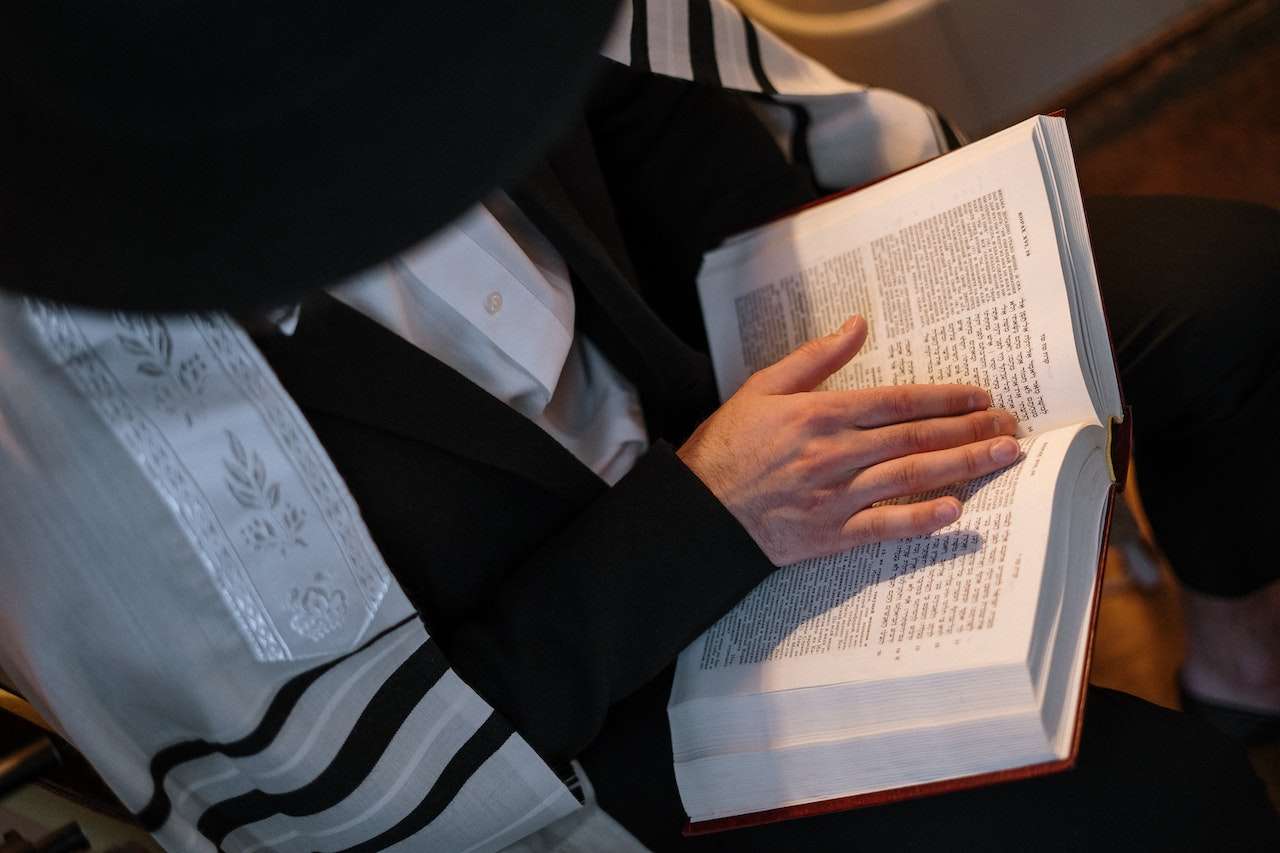 A person sits reading a large open book, possibly a religious text, wearing a white prayer shawl with black stripes and a black hat. Their hand is on the page, and the text is in Hebrew.