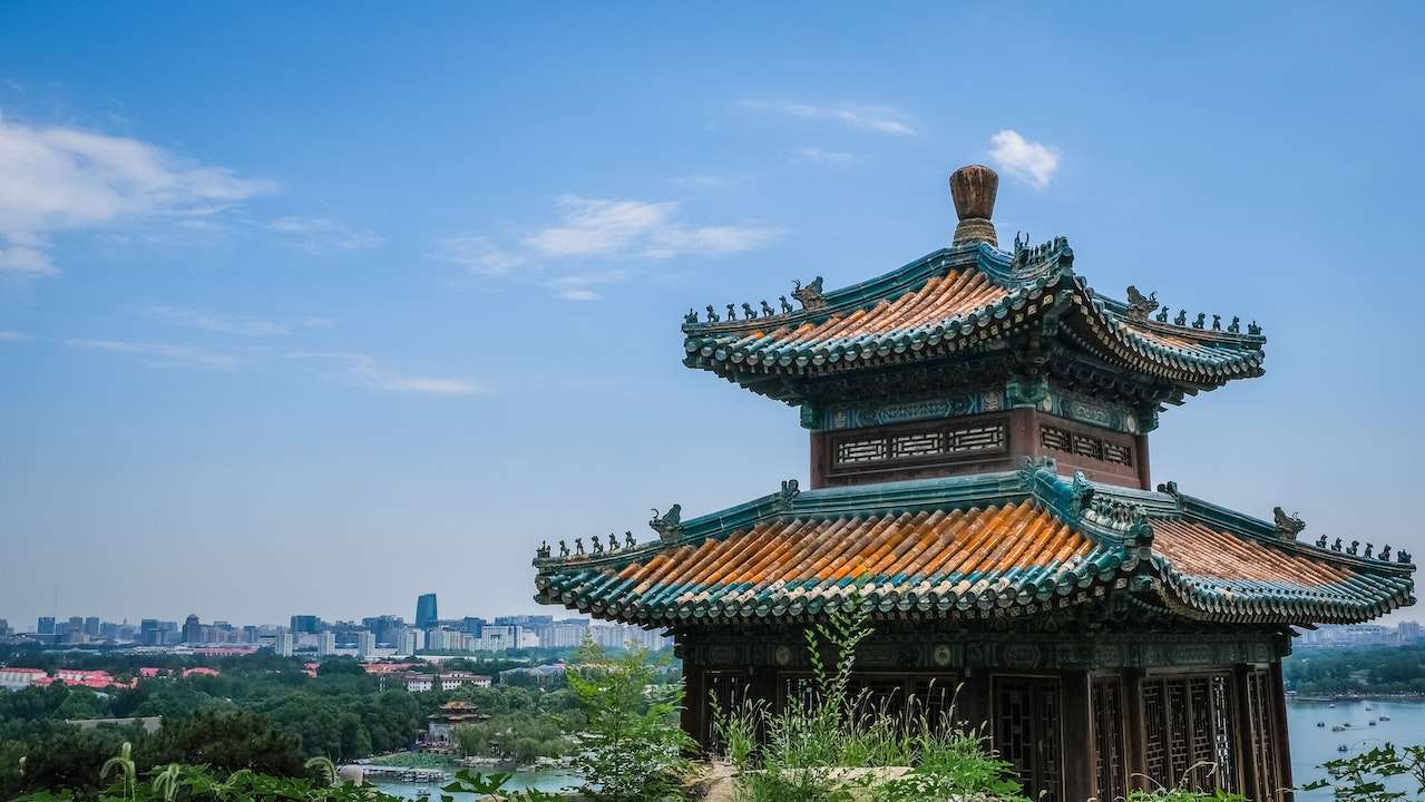 A traditional Chinese pavilion with intricate details and a tiled roof stands in the foreground. In the background, a modern city skyline is visible under a partially cloudy blue sky. Lush greenery surrounds the pavilion, blending nature with architecture.