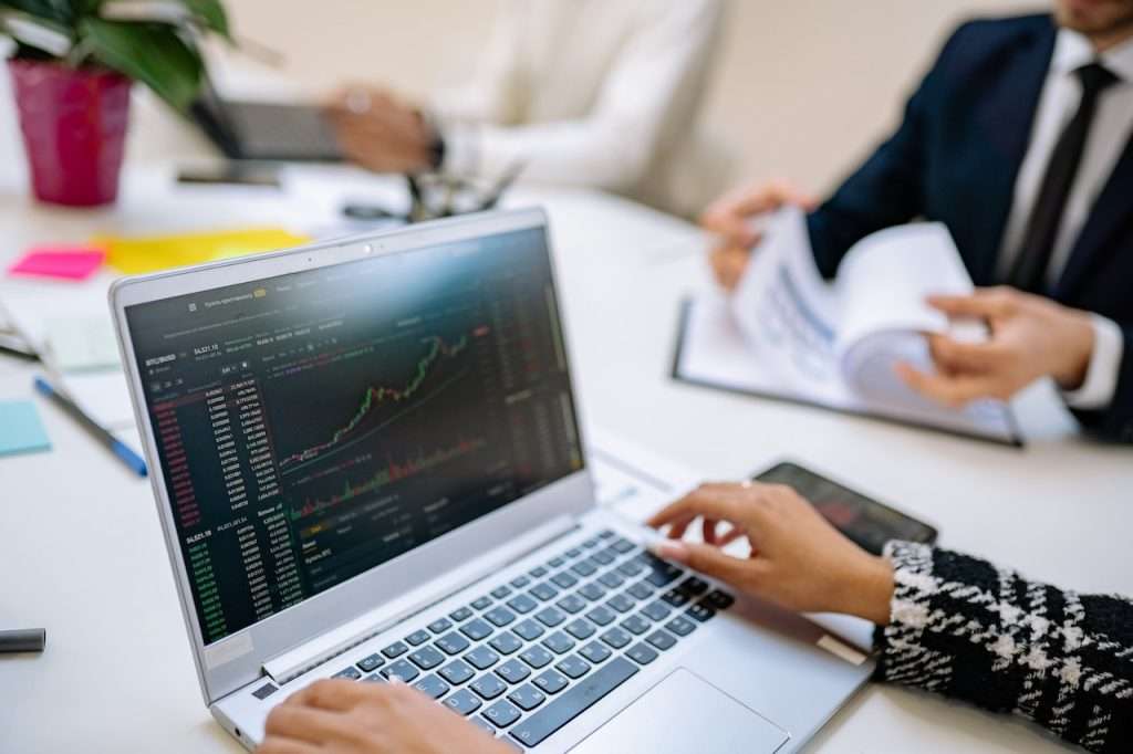 A close-up view of a laptop displaying stock market charts and data. Hands are visible typing on the keyboard. In the background, people in business attire appear to be engaged in a discussion, one holding documents while another operates a tablet.