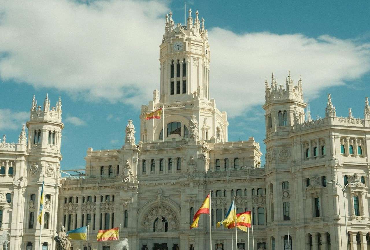 A grand building with intricate architecture and multiple spires, set against a blue sky with scattered clouds. Several flags, including red and yellow ones, can be seen in front of the building. The structure appears historic and majestic.