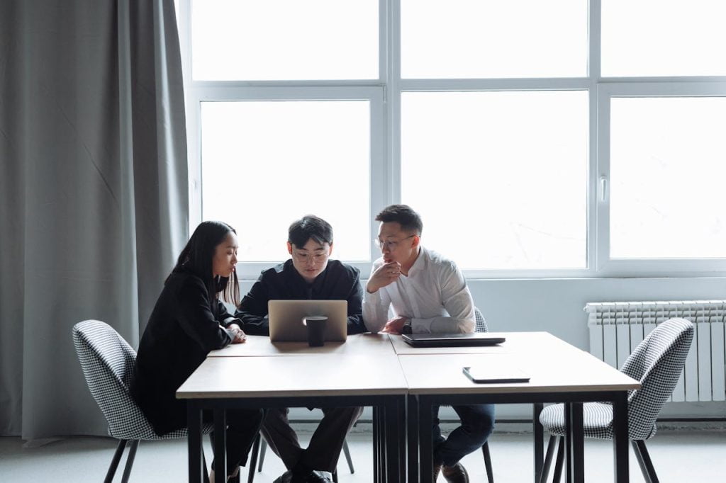 Three people are seated at a conference table in an office, focusing on a laptop screen. A woman and two men appear to be engaged in a discussion. Large windows behind them allow natural light into the room, and the table is mostly clear except for a few items.