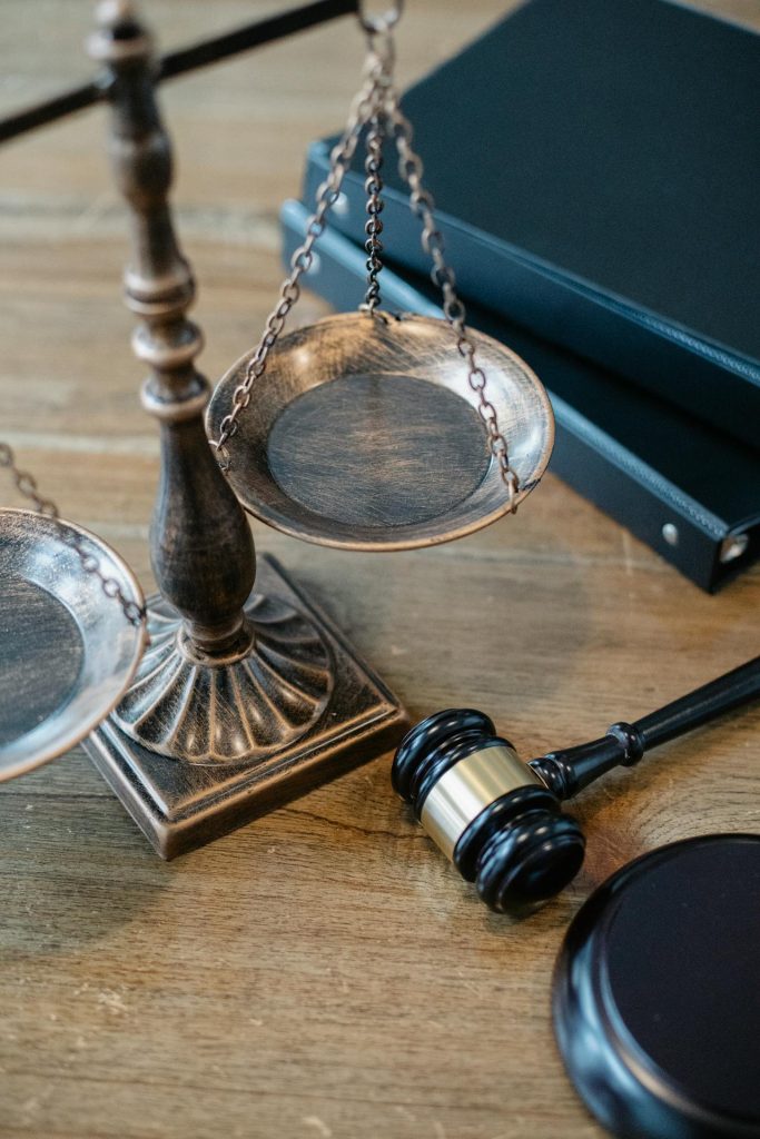 A close-up of a bronze balance scale, a black gavel, and two large black binders on a wooden surface. The balance scale and gavel symbolize justice and the legal system.