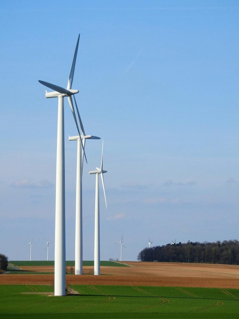 A series of white wind turbines stand tall against a clear blue sky, situated in a lush green field with patches of brown soil. In the distance, more wind turbines are visible, blending into the landscape near a tree-lined horizon.
