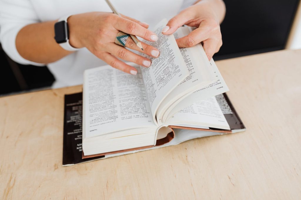 A person, wearing a fitness tracker and a turquoise ring, flips through pages of an open book on a wooden table. The focus is on the hands and the book.