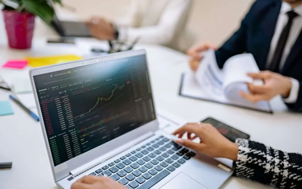 A close-up of a person's hands typing on a laptop displaying a stock market chart. In the background, two people are reviewing documents while seated at a desk. Brightly colored sticky notes and a potted plant are placed on the desk.
