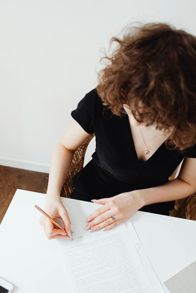 A person with curly hair wearing a black dress is sitting at a white table and writing on a document with a copper-colored pen. The person's head is slightly down, focusing on the paper. A smartphone is placed on the table beside them.