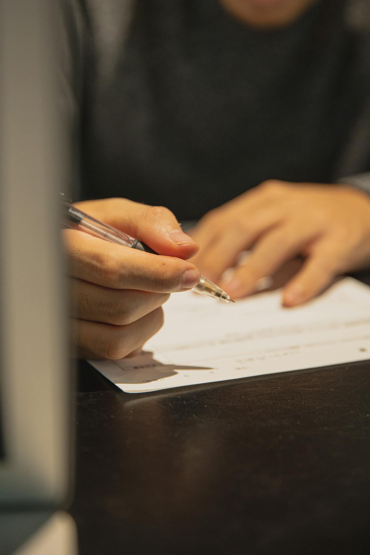 A close-up of a person's hands holding a pen and writing on a sheet of paper. The person is wearing a dark-colored sweater, and a blurred laptop screen is visible in the foreground. The setting appears to be a dimly lit indoor environment.