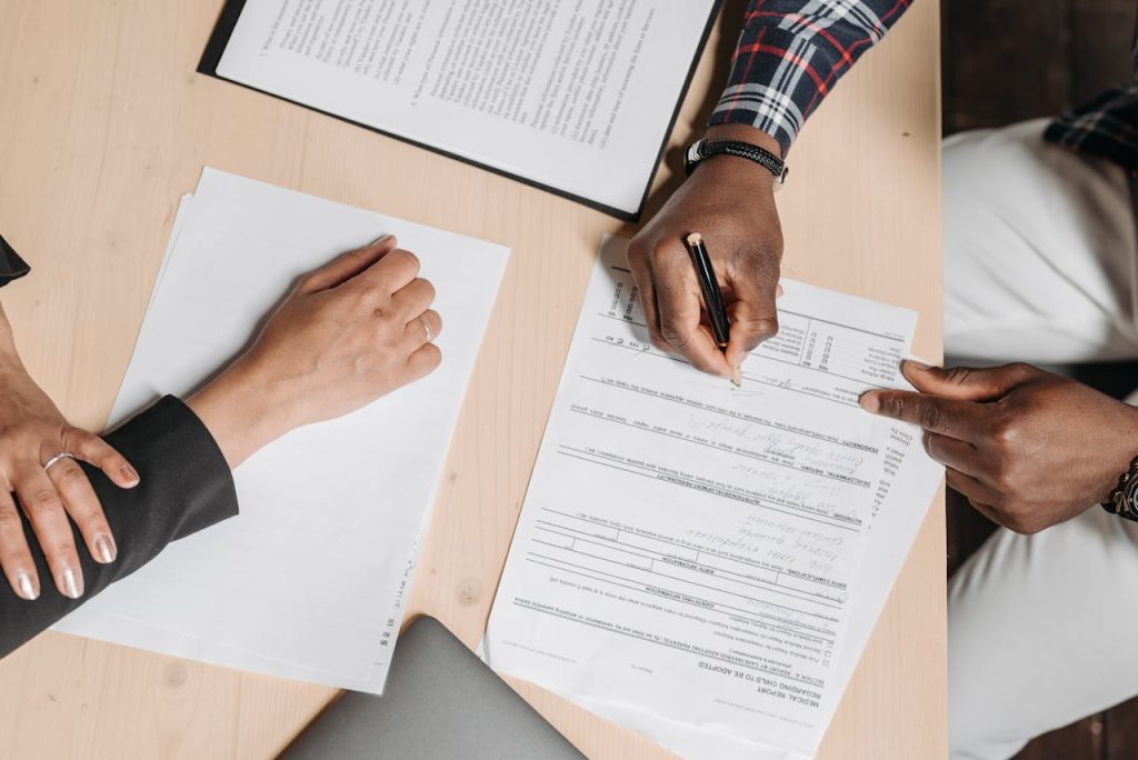 Two people sitting at a wooden desk, reviewing documents. One person points to a section on a form with a pen, while the other person rests their hand on a sheet of paper. A closed folder and a laptop can also be seen on the desk.