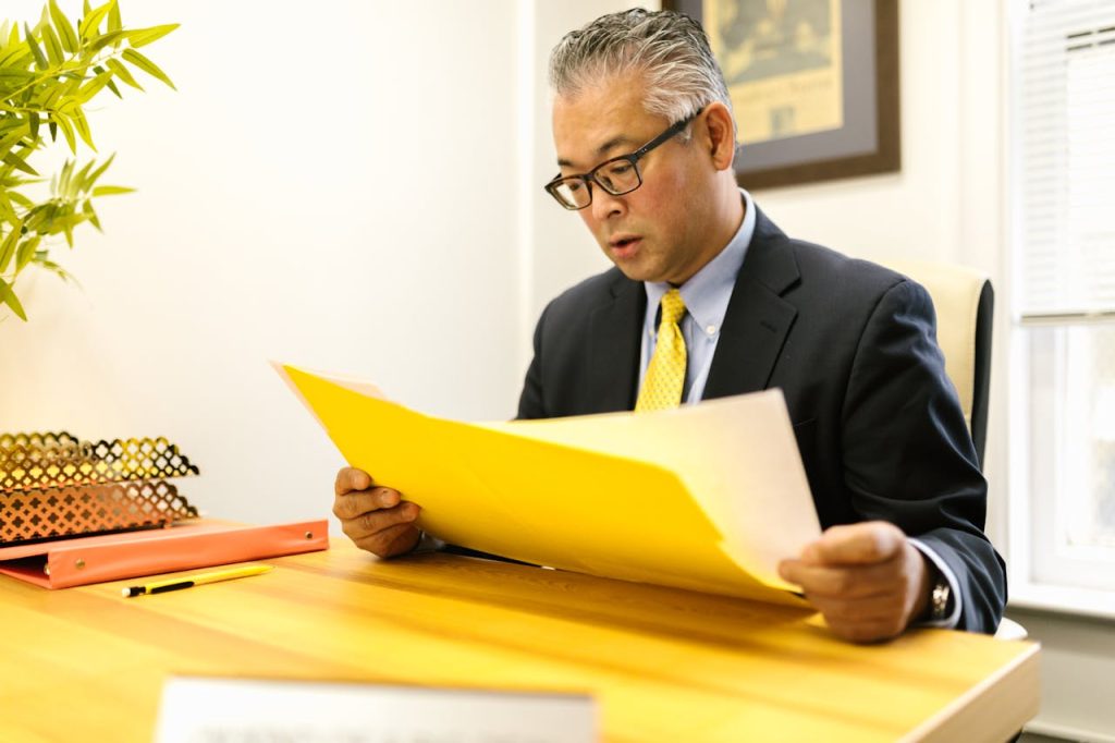 A man in a suit and tie is sitting at a wooden desk, focused on reading a large yellow folder. A green plant, an orange binder, and a pen are visible on the desk. There is a framed picture on the wall behind him and a window to his right.