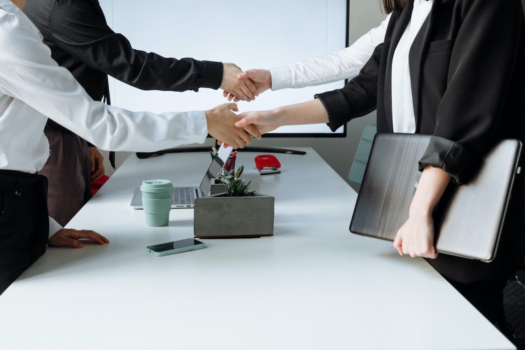 Three people in business attire gathered around a table. Two are shaking hands while standing, and the third person, holding a laptop, stands nearby. A smartphone, coffee cup, and a rectangular planter are visible on the white conference table.