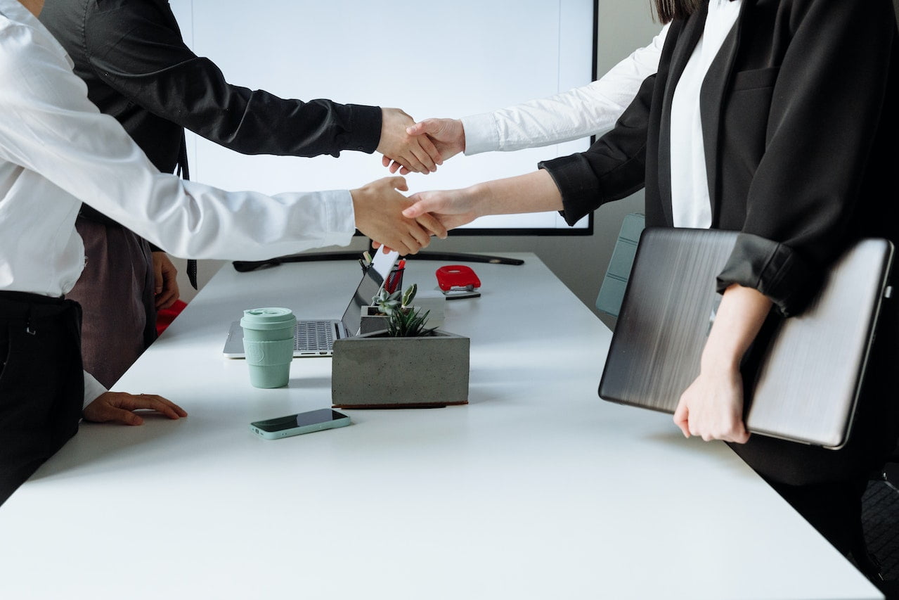 Three people in business attire gathered around a table. Two are shaking hands while standing, and the third person, holding a laptop, stands nearby. A smartphone, coffee cup, and a rectangular planter are visible on the white conference table.
