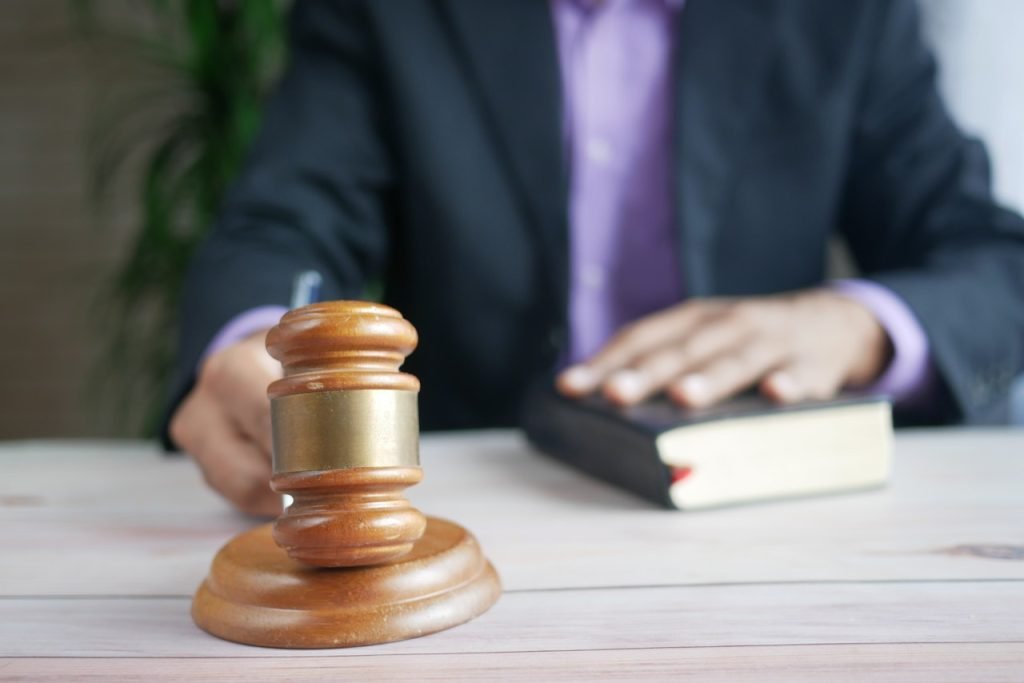 A person in a suit sits at a table with an open book, placing a hand on it, signifying an oath. A wooden judge's gavel is prominently placed in the foreground on the table. The background is blurred.