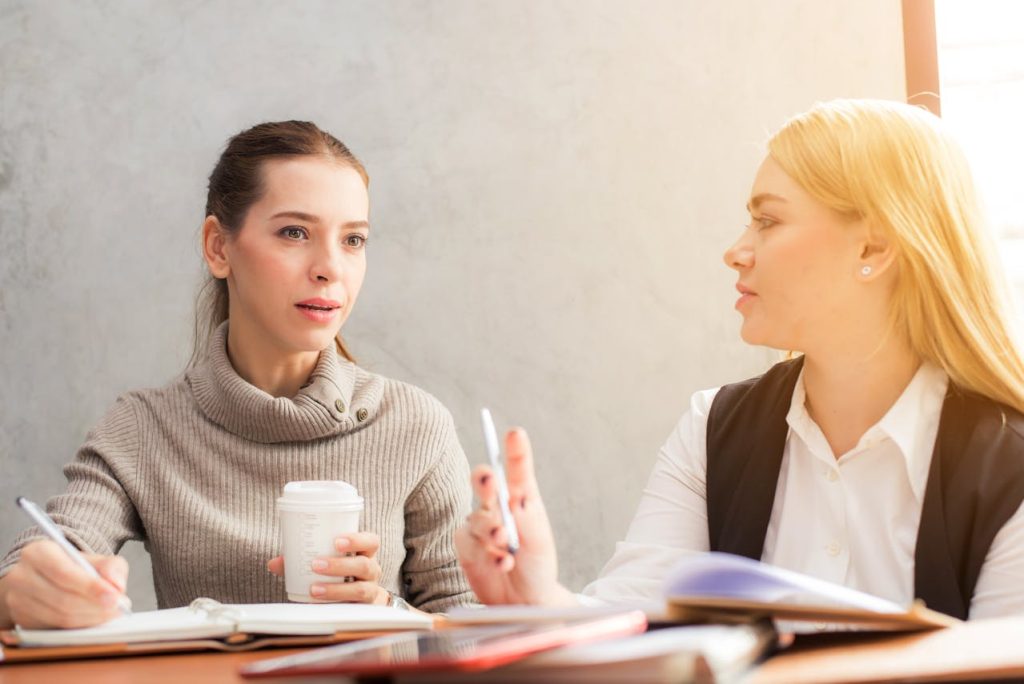 Two women are seated at a table engaged in discussion. One woman, holding a pen and a notebook, appears to be listening attentively, while the other, holding a coffee cup, speaks and gestures with her hand. They are both dressed in professional attire.