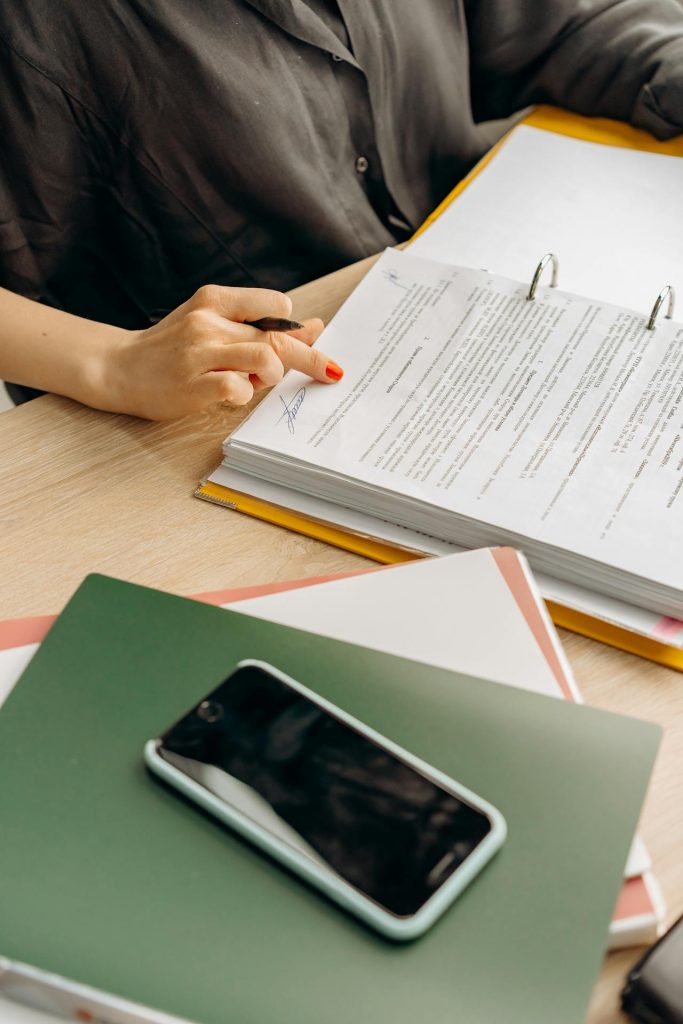 A person wearing a dark shirt sits at a desk, pointing at some documents in a binder with a pen. The desk has a stack of colored folders and a smartphone resting on top. The person has orange nail polish on their fingernails.