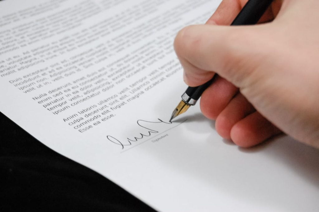 A close-up of a person's hand holding a black fountain pen, signing a document. The paper contains several lines of printed text, and the signature is at the bottom. The background is blurred, making the document and pen the central focus.
