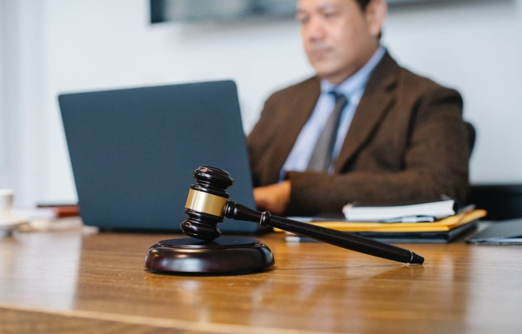 A wooden judge's gavel rests on a desk in the foreground, while a man in a suit works on a laptop in the background, partially out of focus. Papers and a binder are also visible on the desk.