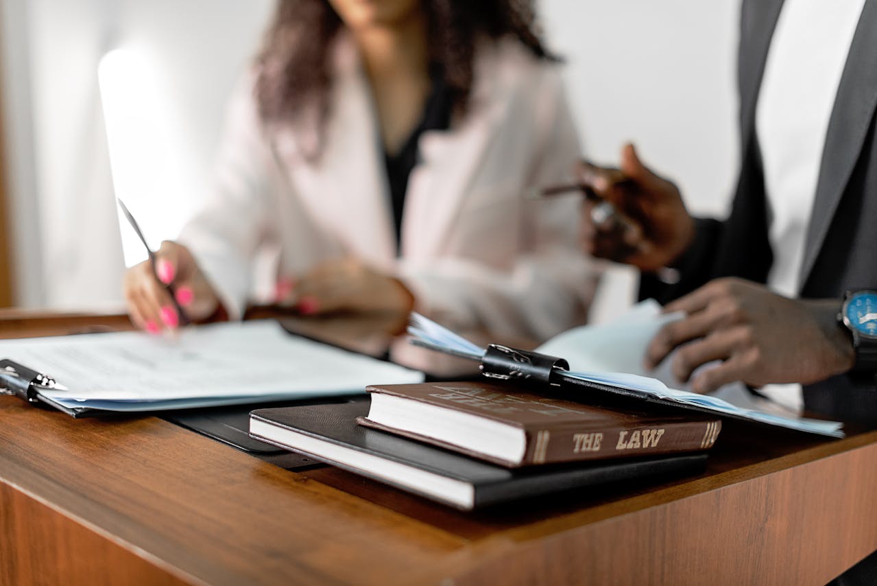 Two individuals are working at a wooden desk. One person is writing on a document, while the other holds a pen, possibly in discussion. In the foreground, there are stacked books, one prominently titled "THE LAW." Their faces are not visible.