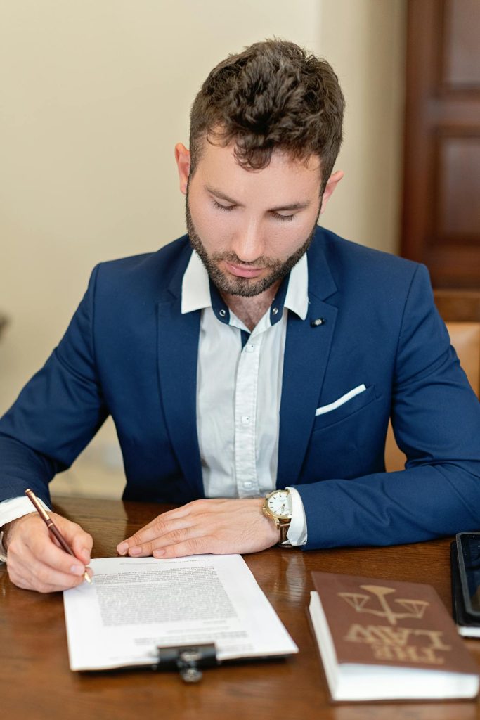 A man in a navy suit with a white shirt is sitting at a desk, focused on writing on a document in front of him. Nearby, there is a book with a legal scales symbol on the cover. The setting suggests a professional or legal environment.